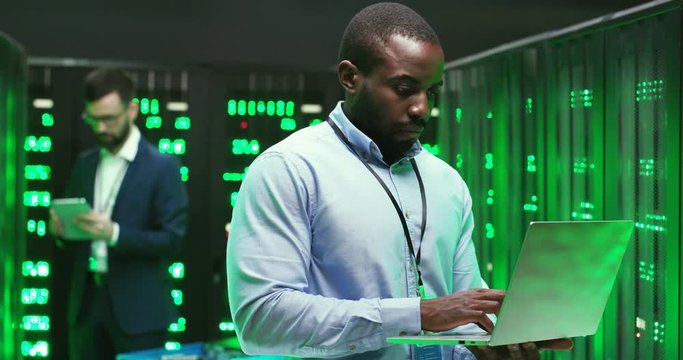 Portrait of handsome African American young man cryptograph standing in big data storage and working as typing on laptop computer. Male Caucasian analytic on background. Information security concept.