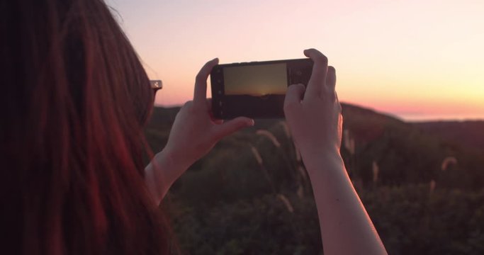 Young Caucasian girl holding phone takes photograph of golden sunset on horizon, Sant'Antioco, Sardinia, Italy, close up pan