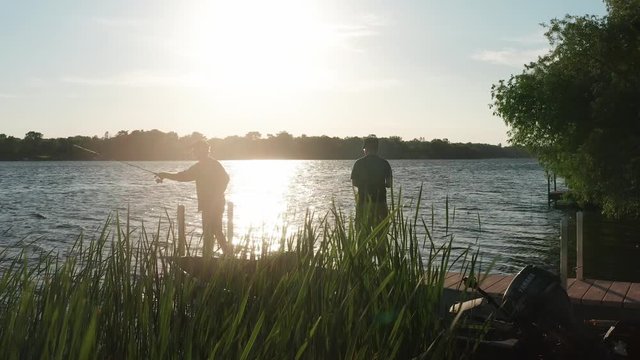 Low shot of father and son fishing on a lake dock at sunset. 4K