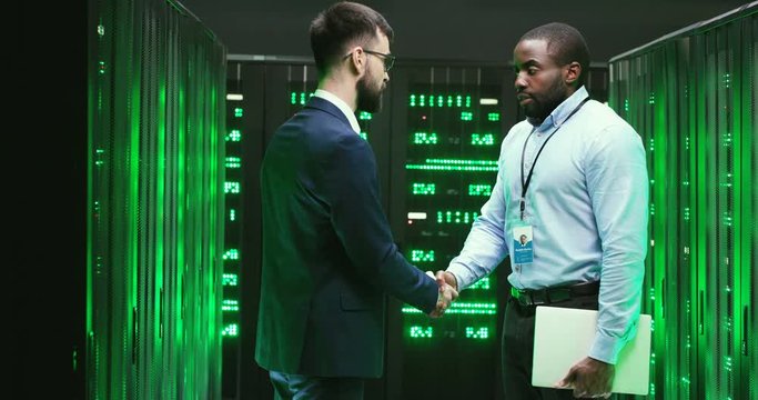 Mixed-races young males engineers standing in digital database of information with servers and talking. Multiethnic men shaking hands and going away. Big data security. Cryptographers.