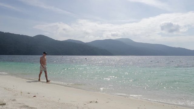 Ultra slow motion shot of young caucasian man with cap and swimwear walking on stunning beach in Thailand