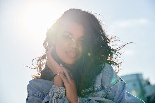 Smiling Beautiful Curly Young Hipster African American Woman Looking At Camera Standing On Sunny Street Backlit With Sunshine. Pretty Gen Z Black Mixed Race Girl Posing For Headshot Portrait Outdoors.