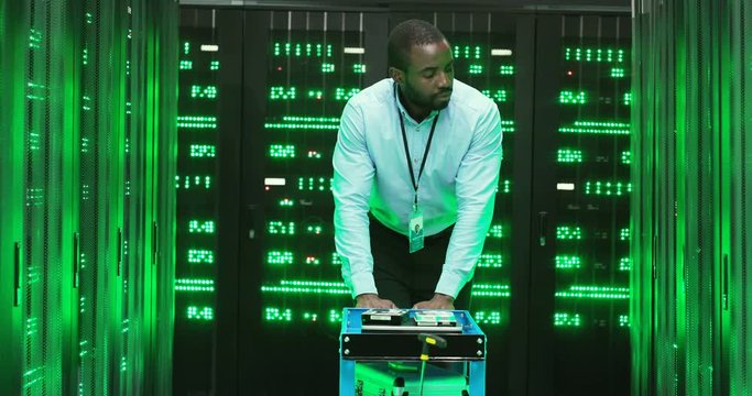 African Amercan handsome man technician walking among servers with big data information and pushing trolley with tools. Green processors in database storage. Male engineer worker of telecommunication.
