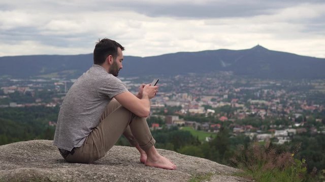 Caucasian man using mobile phone outside, typing and swiping, sitting on rock with awesome view in background