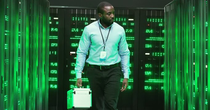 African American man technician with tools box coming in and walking among servers of big data information. Green working processors in database storage. Male engineer worker of telecommunication.