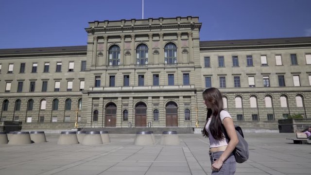 Slow motion mid shot of a female student in grey jeans and pink top with a backpack walking from right to left in front of the university ETH in Zurich