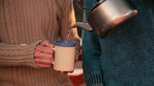 Close up; Man pours hot steaming water from teapot to mug held by woman; sunny outdoors