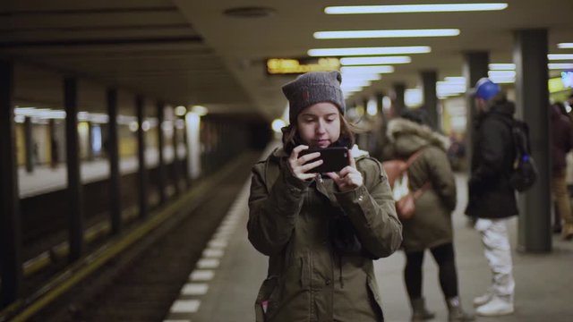 Girl wearing winter hat recording video with her phone at train station platform

