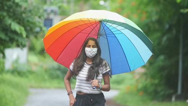 Pretty young dark skinned Indian girl dark hair brown eyes walking towards camera taking mobile phone out of pocket in monsoon rain wearing flowery cotton Mask under a rainbow umbrella slow motion