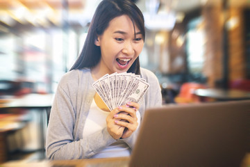 Young woman holding a bunch of dollar banknotes while working online with her laptop. Make money with online business concept.