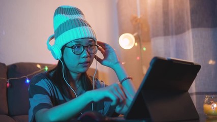 Young hipster looking asian woman wearing headphones and scrolling up and down a tablet touch screen with her finger. Girl browsing music list as entertainment at home. Creative graphic designer - Powered by Adobe