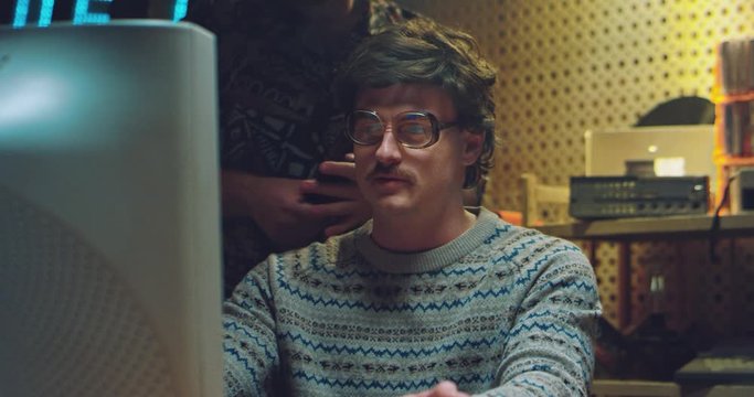 Retro males hackers talking in small room and working on vintage computer. Caucasian man of 80's in glasses and with mustache in front of monitor smoking cigarette. Co-workers technicians of 70's.