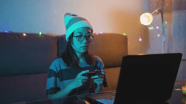 Young adult Asian woman absorbed playing online video games. Girl addited to online gaming. Female gamer using a controller and a laptop at home office