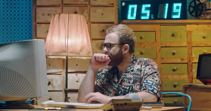 Caucasian young man in glasses and colorful Hawaiian shirt working in vintage room at PC monitor, thinking and typing on keyboard. Male sitting at retro computer. Writer journalists of 80's 90's.