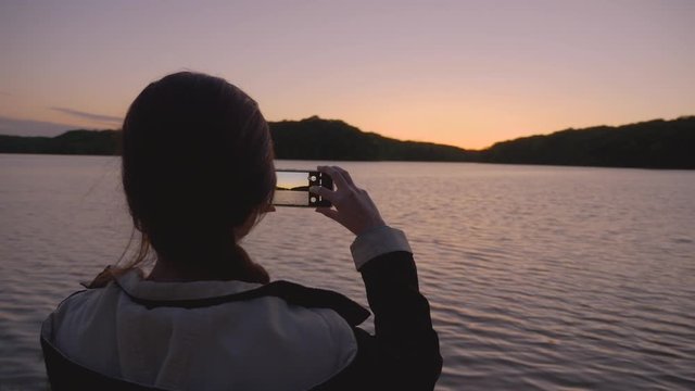 An excited young woman takes a photo of an amazing sunset and lake with her smart phone, slow motion, cinematic, camera spin
