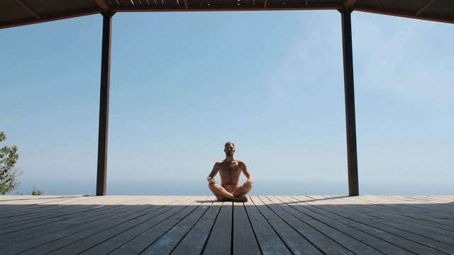 Slow dolly forward footage of a man doing yoga in a special wooden sports house. Handsome muscular bearded man in shorts raises his arms up. Businessman on vacation