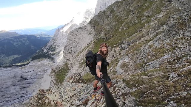 A young, strong and fit man with long hair and tattoos is smiling and stretching out his arm and hand as he reveals the beautiful scenery from the viewpoint on top of the mountain.