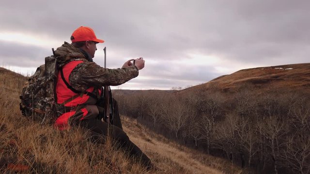 Young Caucasian male hunter with rifle wearing neon orange hat and vest sitting in field taking photos with mobile handheld phone on cloudy day, Saskatchewan, Canada, static close up