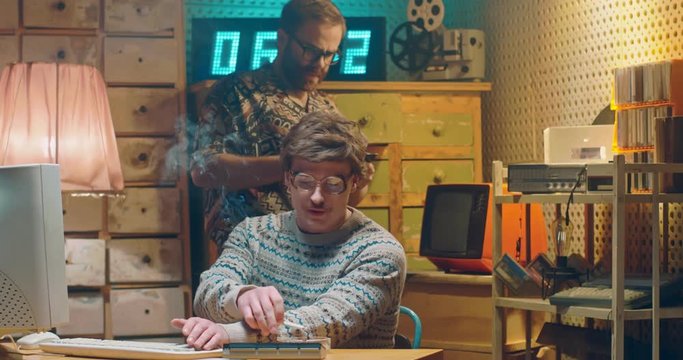 Retro Caucasian males hackers working in vintage interior of room. Man in glasses sitting at old computer and smoking cigarette at desk. Colleague giving diskette with information. Programists of 80's