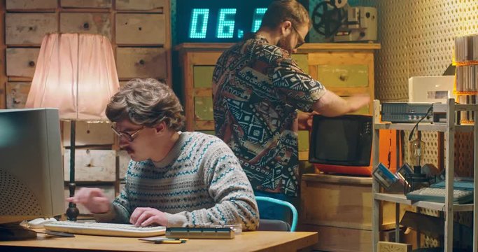 Caucasian nervous computer programist nerd in glasses working at vintage PC monitor and talking with friend. Vintage style room of 90's. Male on background having troubles with old broken TV. Men 80's