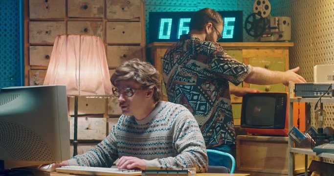 Caucasian computer programist nerd in glasses working at vintage PC monitor and quarreling with friend who disturbing with noise. Vintage style room of 80's. Male on background with old broken TV.