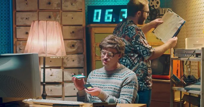 Caucasian young computer programist nerd in glasses sitting at vintage PC monitor and playing with diskettes. Vintage style room of 80's. Male on background with old disk record of 60's or 70's.