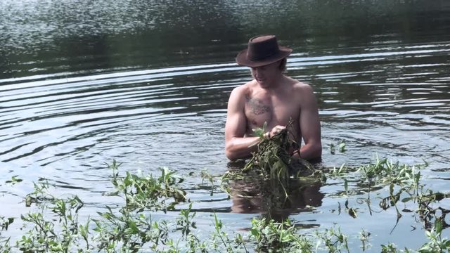 Strong, healthy, shirtless, wet masculine Caucasian adult man wearing outback brown hat standing in lake water pulling tall weeds from pond, handheld close up portrait