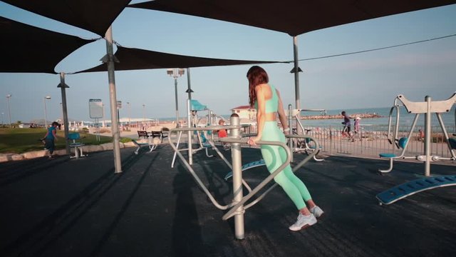 Muscular slim woman lifting herself on parallel bars in an outdoor gym by the beach. Slow motion wide angle shot.