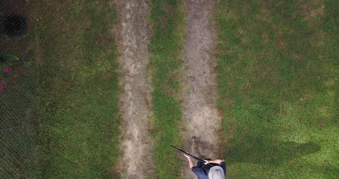Aerial view looking straight down on a man carrying a shotgun in the ready position walking into the frame, stopping halfway he raises the pump action shotgun and cocks it then walks out of frame.