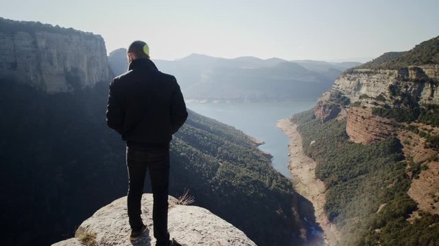 Caucasian man standing dangerously at high edge with sensational scenic views of mountain cliffs and lake water in countryside, handheld pan behind