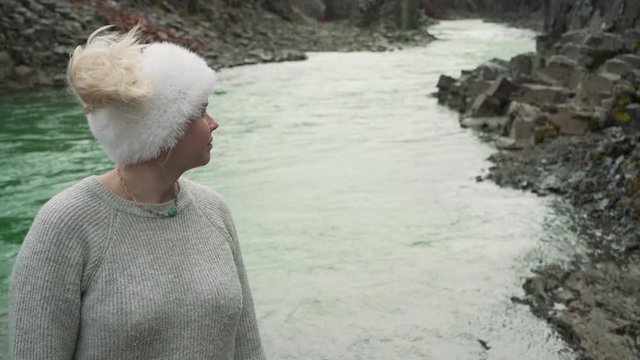 Young Caucasian blonde female wearing a woolen headband standing and starring on the riverbank of an emerald river in Iceland with wind blowing on her.