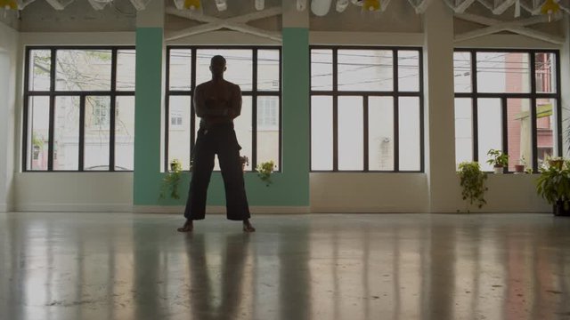 African American black male stands in empty studio wearing black karate pants, shirtless, performing karate kias. Wide frame, 4K, windows in BG.