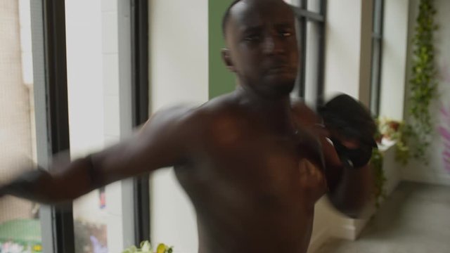 African American black male wearing black karate pants, shirtless throws fast kicks and punches with boxing gloves at lens. Wide medium frame, 4K, gimbal, interior studio.