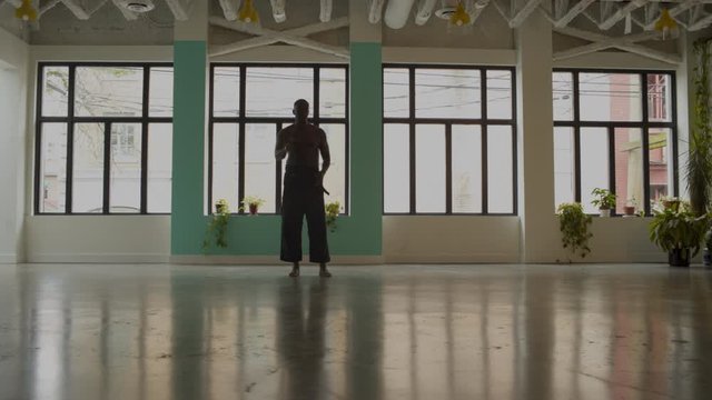 African American black male wearing black karate pants, shirtless, in fitness studio with windows skipping. 4K wide frame.