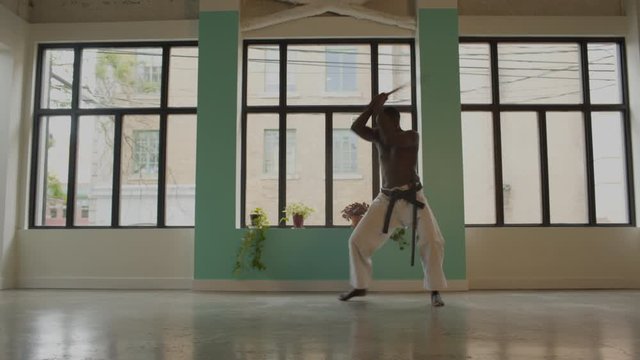 African American black male wearing karate pants, shirtless, in an empty studio with windows, moves through a fast black belt sword routine from kneeling to standing. 4K wide frame.