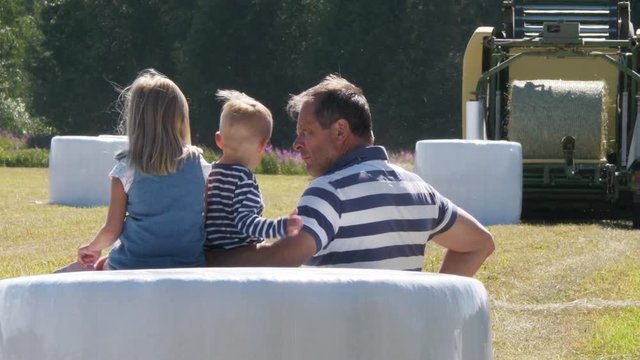 Farmer and Grandchildren Watching Hay Bale Roll Being Wrapped in Plastic For Protection