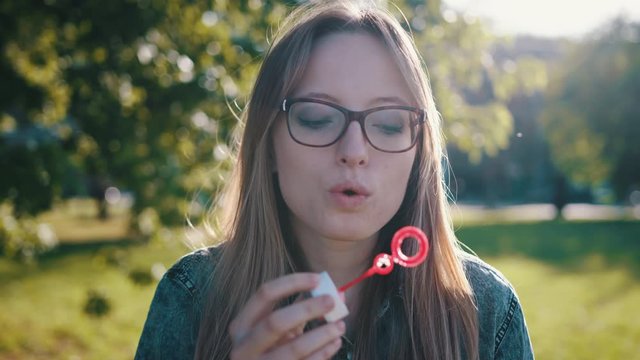 Young woman blowing soap bubbles in nature. A happy young brunette girl with glasses in the park