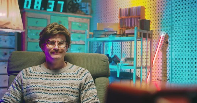Caucasian weeping young man nerd in glasses and with mustache sitting in retro room and watching TV with tears. Vintage television of 80's. Male crying at sad drama movie. Goofy guy of 70's Zooming in