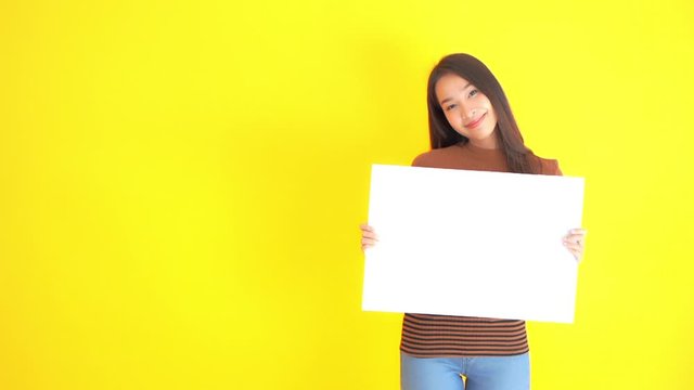 Young natural asian woman holding empty white board, for logo, slogan or message slow motion, isolated on yellow background, full frame