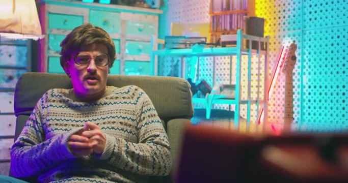 Caucasian afraid young man nerd in glasses and with mustache sitting in retro room and watching film on TV. Vintage television of 80's. Male is scared of horror movie. Goofy guy of 70's Scare and fear