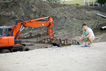 A young father and son on a construction site. They look at the excavator.