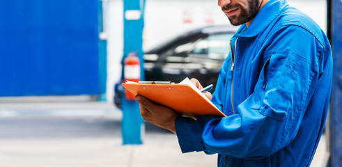 Crop image of mechanic in blue work wear uniform checks the vehicle maintenance checklist with blur lifted car in the background. Automobile repairing service, Professional occupation.