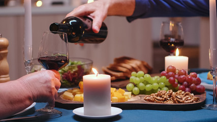 Close up of retired senior man pouring red wine in wife glass. Romantic retired old couple celebratic at candle lights, love and anniversary