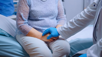 Close up of doctor hands in blue protective gloves comfortin a stressed old woman sitting on hospital bed in modern private clinic. Global pandemic coronavirus covid-19 healthcare pandemic