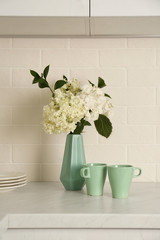 Bouquet with beautiful white hydrangea flowers and cups on light countertop