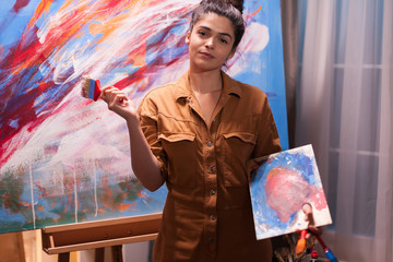 Portrait of young painter in art studio holding paint brush. Modern artwork paint on canvas,...