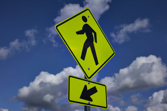 Pedestrian crossing sign against blue sky. A yellow warning sign that alerts road users to unexpected conditions ahead and reminds them that it is a place designated for pedestrians to cross a road. - Powered by Adobe