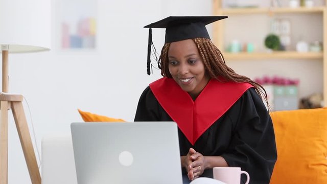 woman sit on sofa in cap and gown and talk to relatives on video laptop call Spbi. proud african american female college graduating. certificate, achievement, discussion concept
