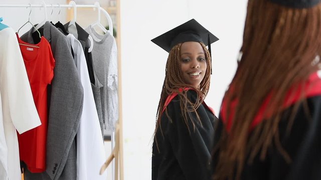 african woman trying on, fitting cap and gown and looking in mirror with happy smile Spbi. put on graduating clothes in wardrobe. concept graduation, clothes, american