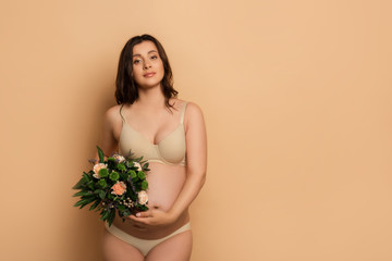  pregnant woman in lingerie holding bouquet while looking at camera on beige